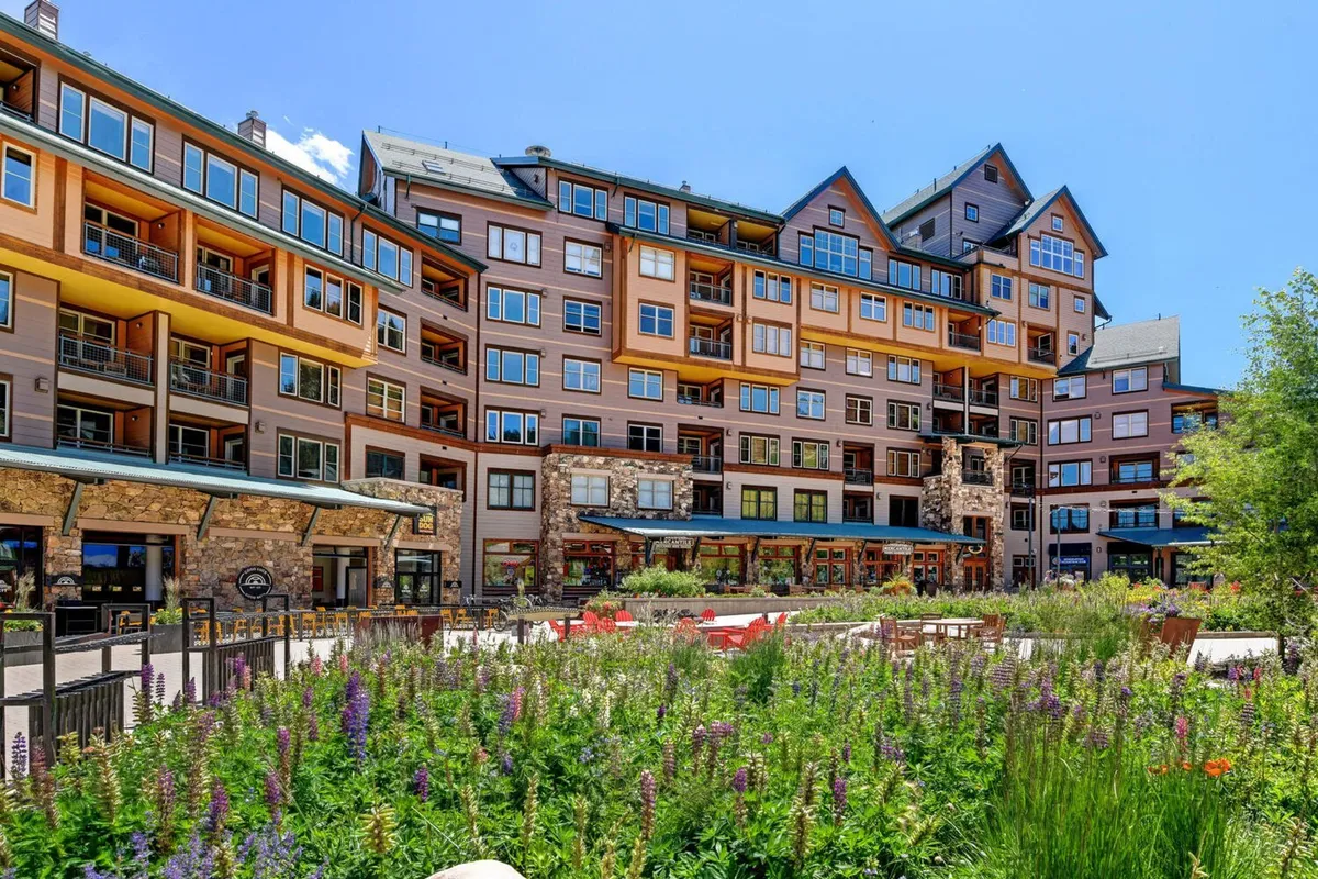 A vibrant exterior scene featuring a multi-story building with colorful flower beds and an inviting outdoor seating area. Groups of people are relaxing and sharing meals, while others stroll through the lively garden setting. The overall mood is welcoming and energetic, suggesting both daytime and evening gatherings.