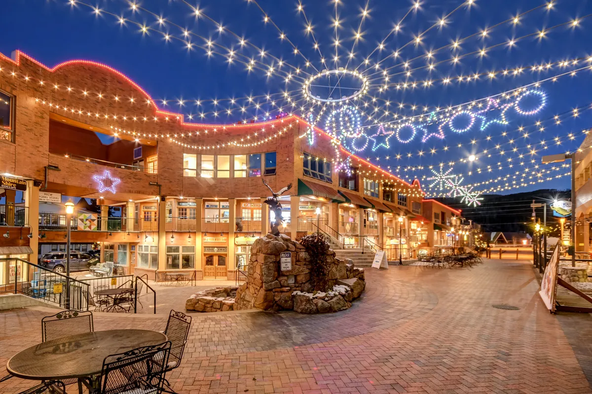 A magical nighttime exterior view with strings of lights illuminating a decorative archway, outdoor seating, and a stone fountain. The area is bustling with people socializing, dining, shopping, and enjoying the festive atmosphere of the village at night.
