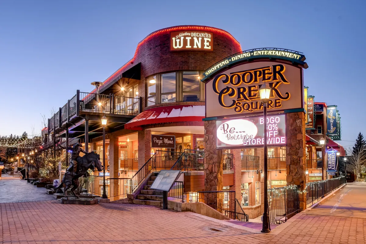 A bustling village square, marked by the Cooper Creek Square sign, features outdoor seating areas, decorative lighting, and signs for local wine shops. The scene is lively, illustrating guests enjoying the social energy, shopping, and entertainment that the location offers.