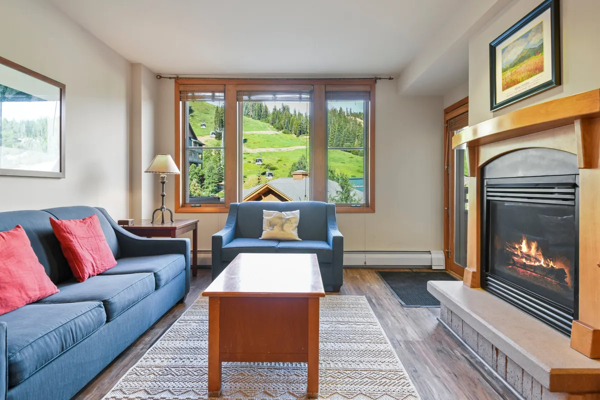 A cozy lobby lounge area featuring a grand stone fireplace, a comfortable sofa, and large windows that frame views of the snowy outdoors. The ambiance is warm and inviting, perfect for gathering or relaxing after time spent on the slopes.