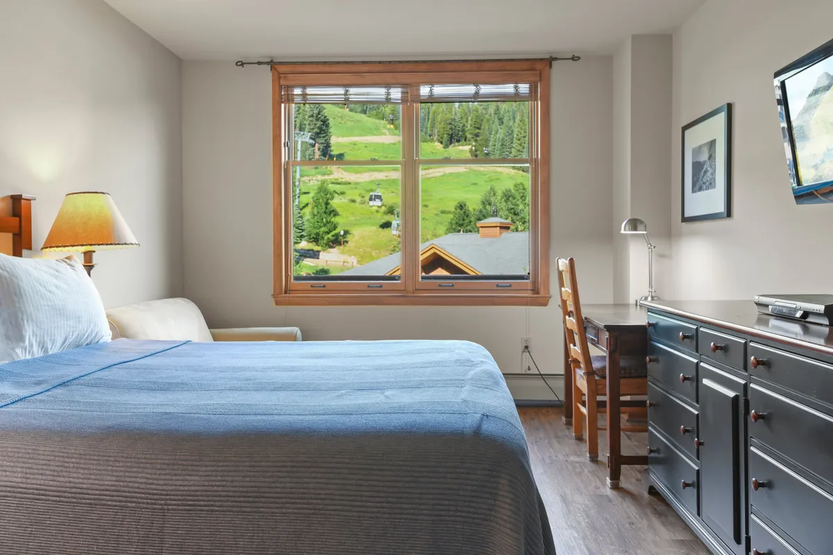 The image shows a bed with a blue blanket, a large window opening to mountain views, a wooden desk with a chair, and a bedside lamp. The decor is tranquil, and the natural surroundings enhance the sense of retreat and restfulness.