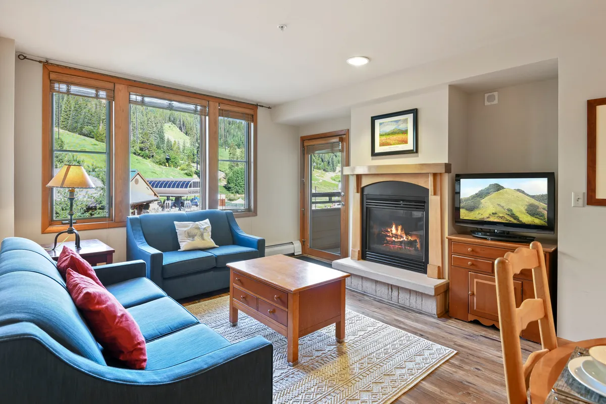 The image shows a cozy, inviting living room with a glowing fireplace as the focal point, plush sofa and coffee table arranged for conversation or games, and expansive windows offering stunning mountain views. The atmosphere is warm and welcoming, perfect for relaxing after a day of adventure.