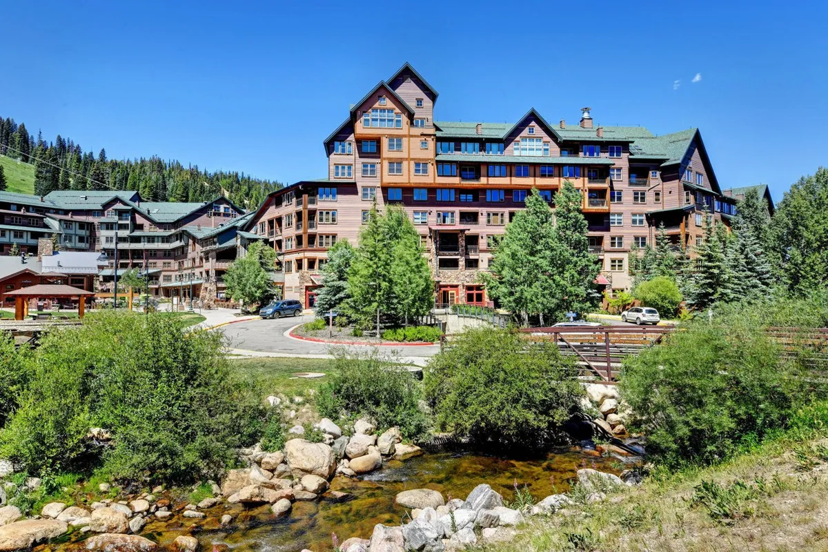 This exterior image captures the main building surrounded by lush trees, a flowing stream, and a winding pathway. The natural landscape hints at adventure and exploration, while the proximity of the building to these outdoor elements emphasizes easy access to nature and activities.