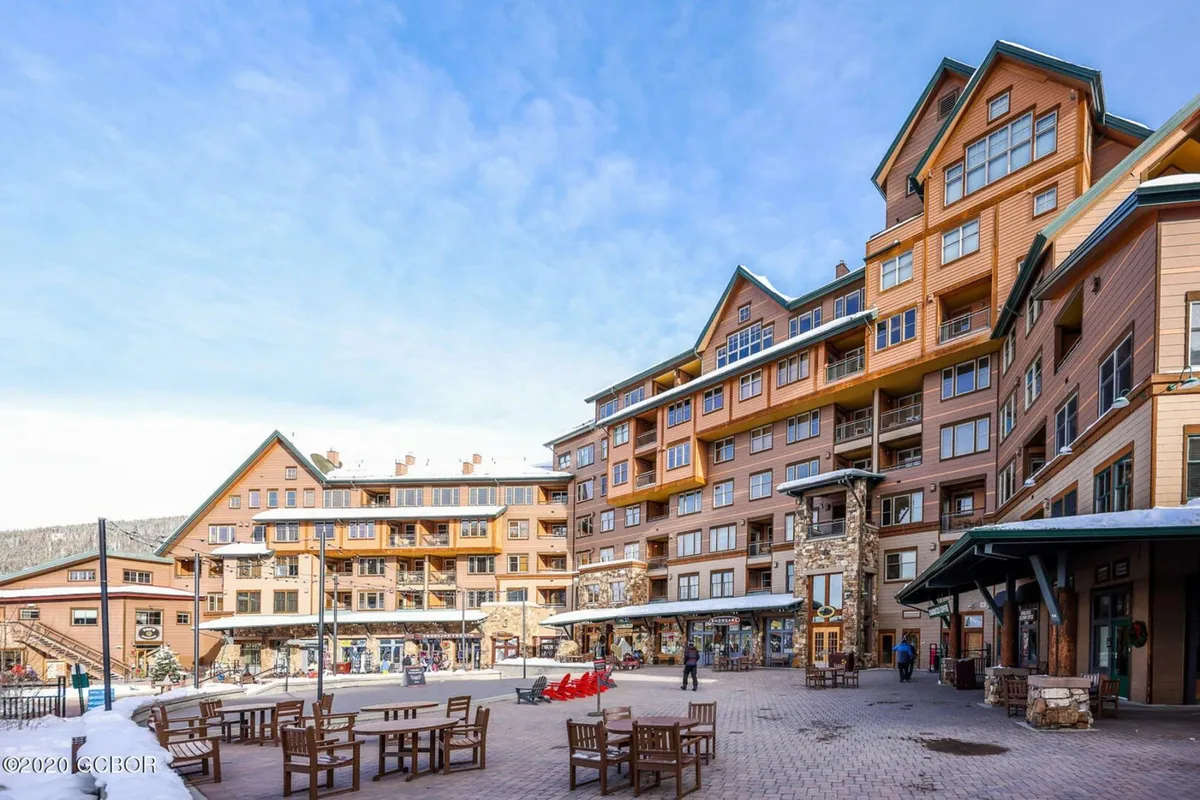 Capturing a vibrant plaza with outdoor seating, people enjoying the fresh air, and a multi-story building with a clock tower, this image conveys the lively energy of the Winter Park Resort village below the deck. Red chairs and people walking create a cheerful atmosphere, emphasizing community and activity.
