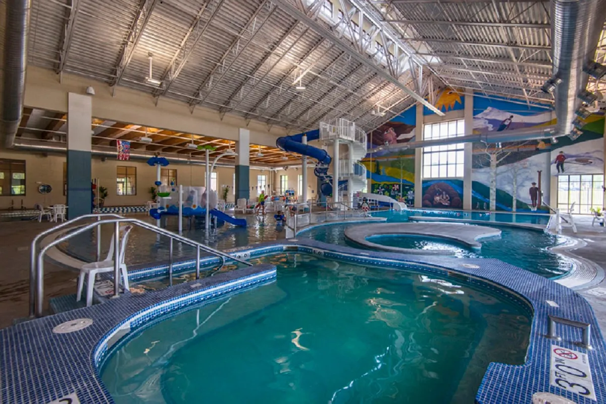 A vibrant interior shot of the local recreation center's pool area, where children and families play on colorful water slides and in the pool. The atmosphere is lively and energetic, featuring bright murals and a sense of fun.