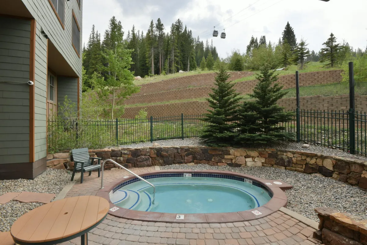 An inviting exterior hot tub set among surrounding trees with a backdrop of mountains. The hot tub area features a fence and outdoor seating, with the warm water reflecting the soft colors of sunset, suggesting a peaceful setting for après-ski relaxation.