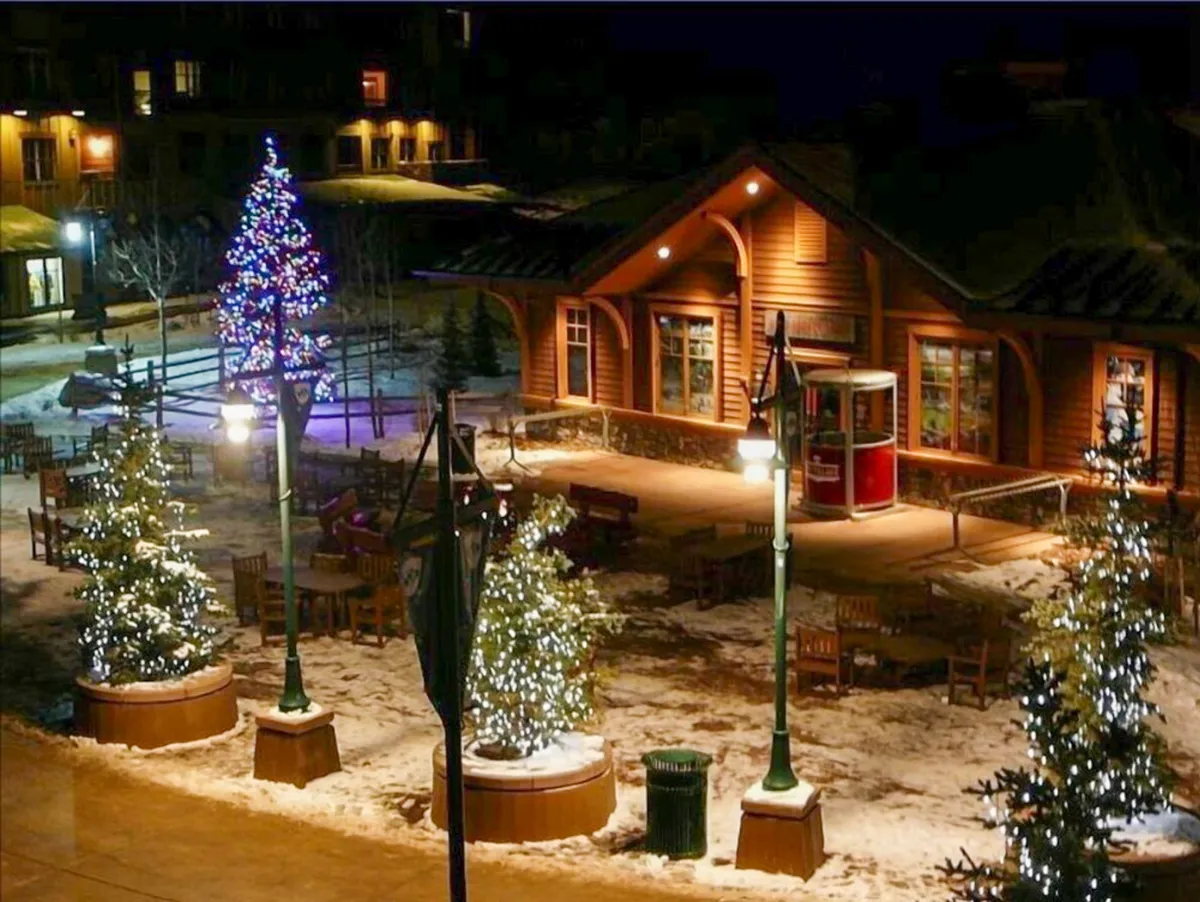 This image captures an exterior winter scene with a beautifully decorated Christmas tree, illuminated building, lighted shrubs, and an inviting outdoor seating area. People can be seen gathering, celebrating, and enjoying the festive ambiance of the alpine village at night.