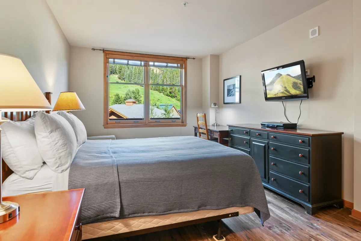 This image features a serene bedroom with a bed, a window offering a pleasing view, a television, a dresser, and a desk. The space is bathed in natural light and styled for both relaxation and functionality, evoking a tranquil and comfortable atmosphere.