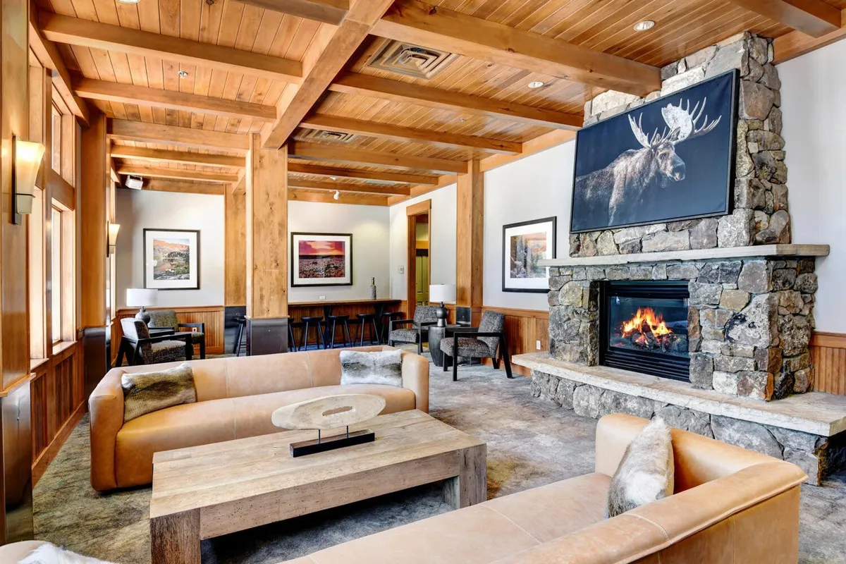 This inviting living room features a glowing gas fireplace as its focal point, complemented by large moose artwork above the mantle, exposed wooden beams, and a plush, comfortable seating arrangement. The ambiance is warm and welcoming, designed for gathering and relaxation after a day of mountain adventure.